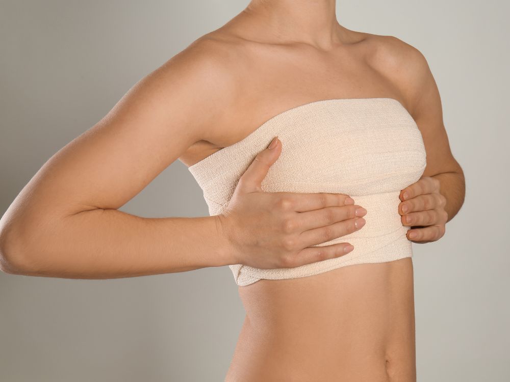 Mulher com bandagem nos seios após cirurgia. A dor na mama após mamoplastia redutora pode ocorrer devido à cicatrização e inflamação no pós-operatório.