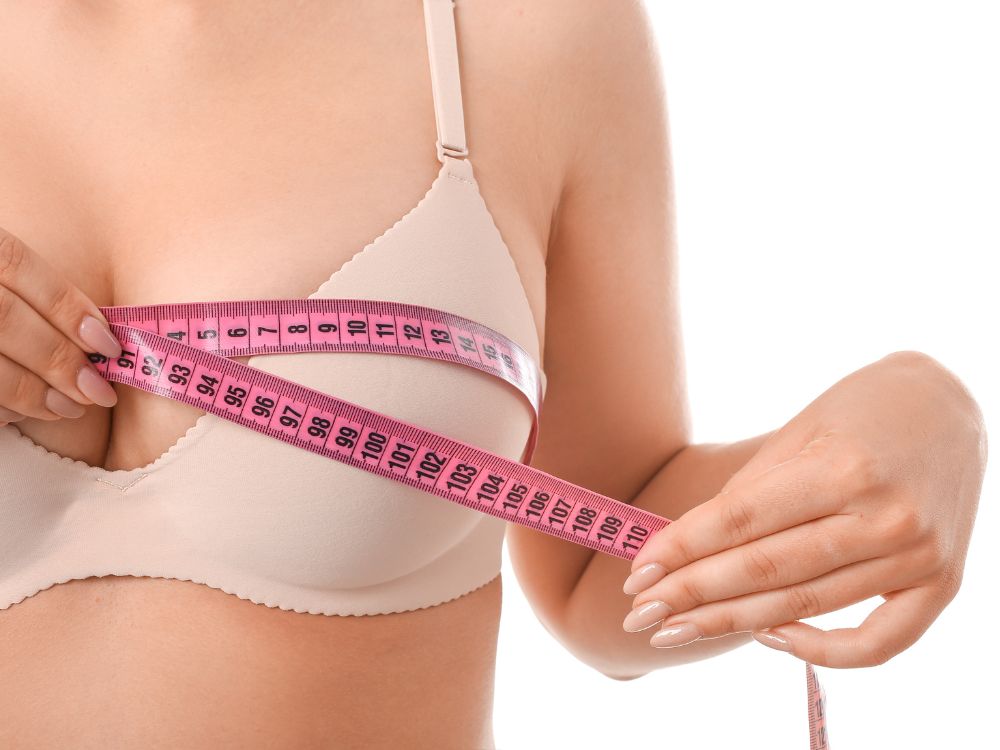 Mulher medindo o tamanho dos seios com fita métrica antes da cirurgia. Entenda como a mamoplastia redutora risco de morte pode ser minimizado.