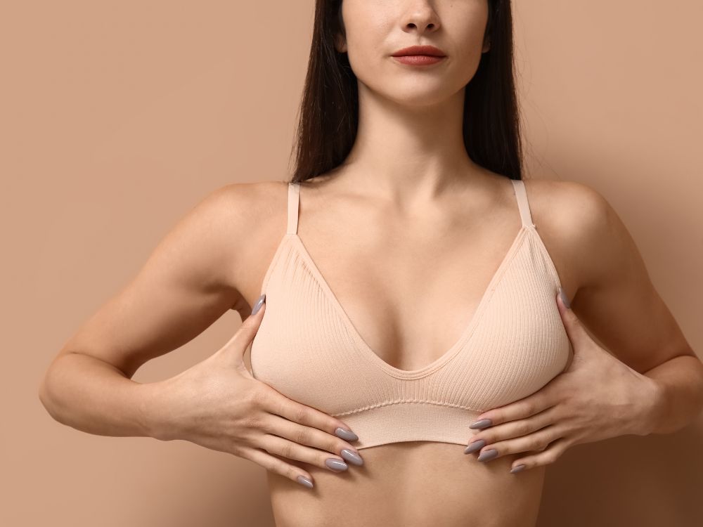 Mulher com sutiã bege segurando os seios para ilustrar cuidados e resultados de o que é mamoplastia.
