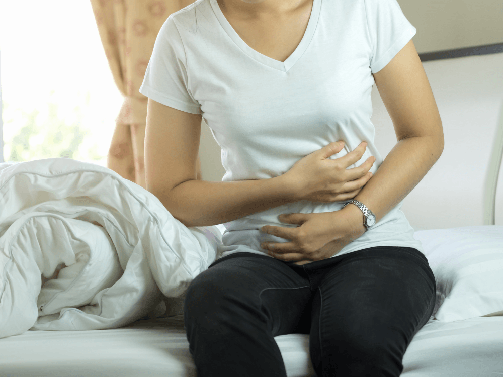 Paciente sentada na cama segura o abdômen durante recuperação, situação comum nos primeiros dias após cirurgia.