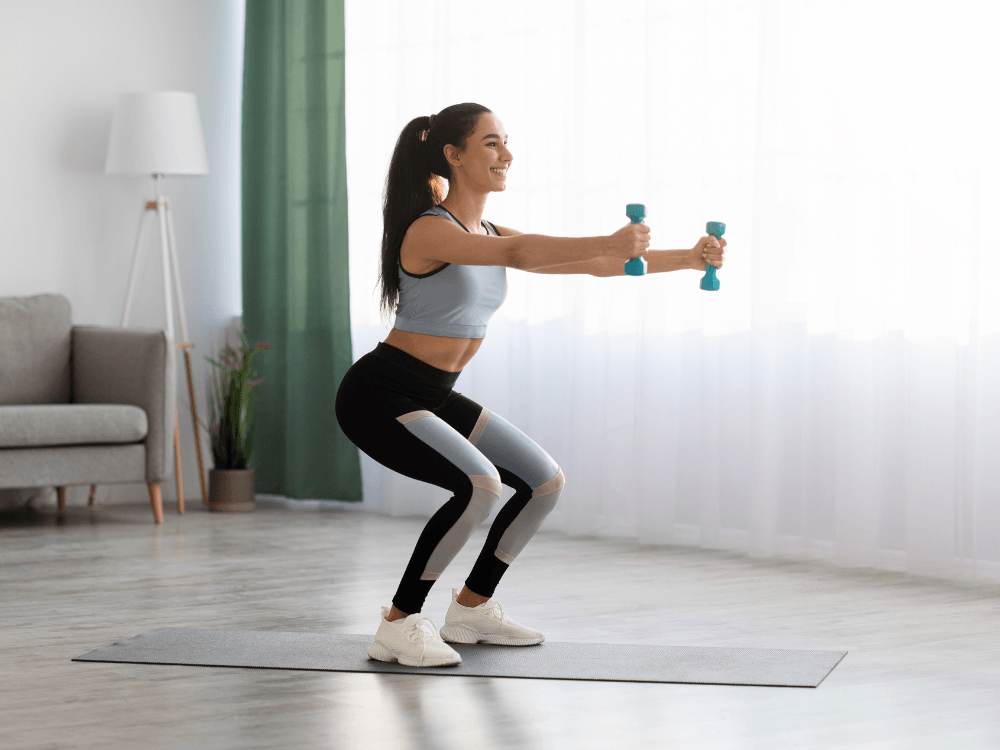 Mulher realiza agachamento com halteres em casa, exercício que exige atenção ao retorno gradual após uma cirurgia abdominal.