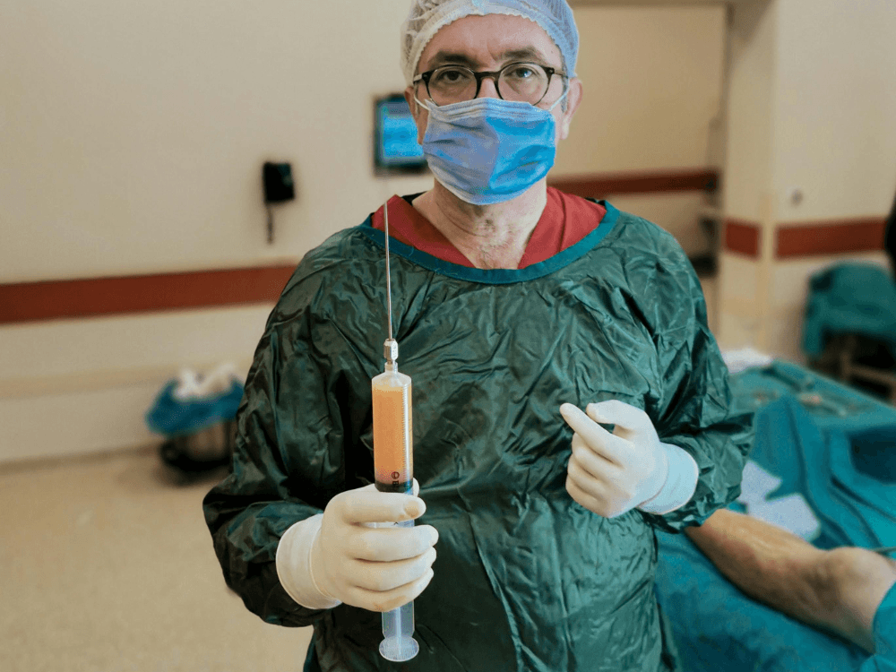 Cirurgião plástico paramentado segura uma seringa com líquido durante procedimento em ambiente hospitalar de cirurgia estética.